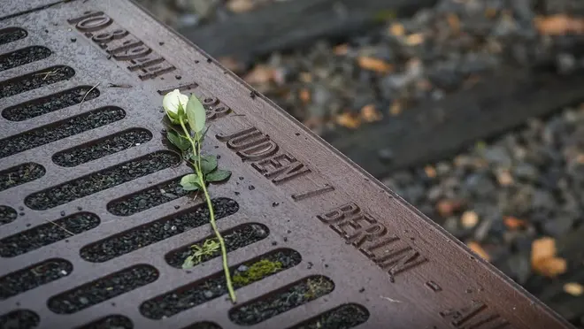 Einladungscover der Gedenkveranstaltung "abgeholt" am 18. Oktober 2026 mit einem Farbfoto: auf dem Mahnmal liegt eine weiße Rose. Im Hintergrund sind Gleise zu sehen.