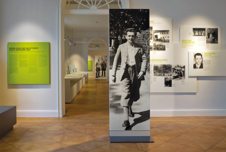 Blick in die Dauerausstellung: Eine etwa zwei Meter hohe Stele zeigt eine Schwarz-Weiß-Aufnahme von Georg Elser in Jackett und Kniebundhose. Im hinteren Bereich der Ausstellung steht eine Stele mit einer Schwarz-Weiß-Aufnahme von Claus Schenk Graf von Stauffenberg und Albrecht Ritter Mertz von Quirnheim in Militäruniform.