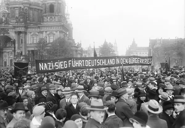 Historisches Schwarz-Weiß-Foto: Eine Menschenmenge steht dicht zusammen. Menschen halten Fahnen und Plakate hoch. Auf einem Banner steht: Nazi-Sieg führt Deutschland in den Bürgerkrieg.