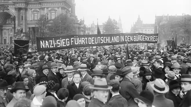 Historisches Schwarz-Weiß-Foto: Eine Menschenmenge steht dicht zusammen. Menschen halten Fahnen und Plakate hoch. Auf einem Banner steht: Nazi-Sieg führt Deutschland in den Bürgerkrieg.