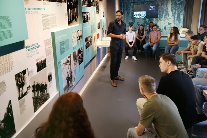 Ein Referent steht vor einer Ausstellungswand, ihm gegenüber sitzen Schülerinnen und Schüler auf einer Bank und hören ihm zu.